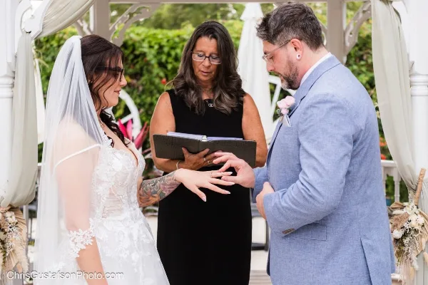 A wedding officiant in a black dress officiating a ceremony and placing a ring on the bride's finger.