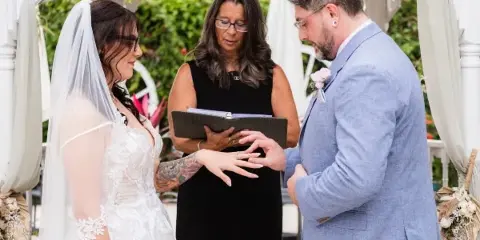 A wedding officiant in a black dress officiating a ceremony and placing a ring on the bride's finger.