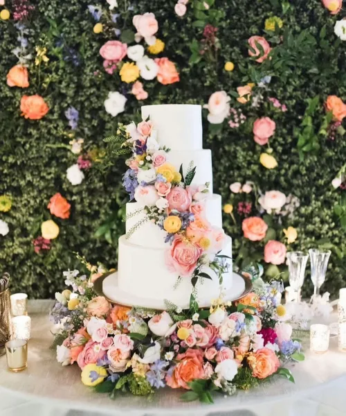 A white tiered wedding cake decorated with pink and yellow flowers, set against a lush green floral wall backdrop.