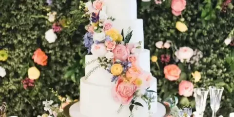 A white tiered wedding cake decorated with pink and yellow flowers, set against a lush green floral wall backdrop.