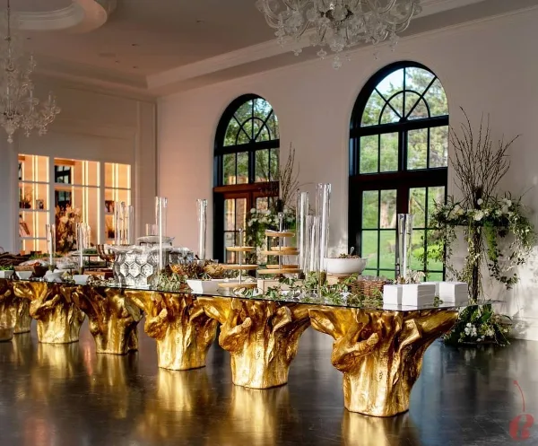 A long glass event table with gold sculptural bases, decorated with floral arrangements and glassware in a room with arched windows and chandeliers.