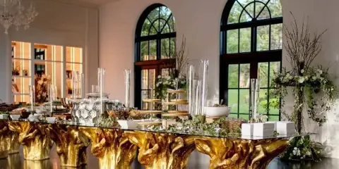 A long glass event table with gold sculptural bases, decorated with floral arrangements and glassware in a room with arched windows and chandeliers.