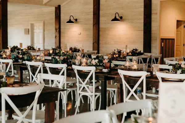 Interior view of a rustic wedding venue reception hall with white wooden walls, dark timber beams, and long tables set with white chairs and floral centerpieces.