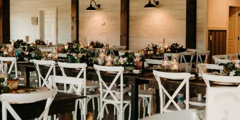 Interior view of a rustic wedding venue reception hall with white wooden walls, dark timber beams, and long tables set with white chairs and floral centerpieces.