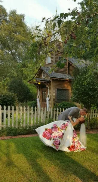 A bride and groom in a wedding pose in front of a rustic wooden chapel and white picket fence at a countryside venue.