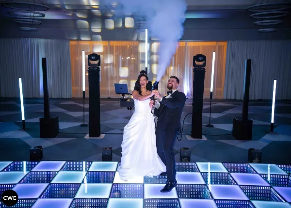 A bride and groom on an LED dance floor with stage lighting, speakers, and a CO2 cold spark effect at a wedding event.