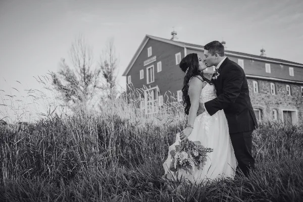 A black and white photograph of a bride and groom kissing in a field of tall grass with the Kleffner Ranch barn in the background.