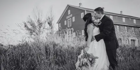 A black and white photograph of a bride and groom kissing in a field of tall grass with the Kleffner Ranch barn in the background.