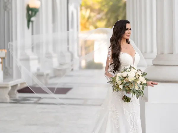 A bride with long wavy hair and a veil holding a bouquet of white flowers in a white wedding dress, standing in a colonnade.