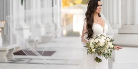 A bride with long wavy hair and a veil holding a bouquet of white flowers in a white wedding dress, standing in a colonnade.