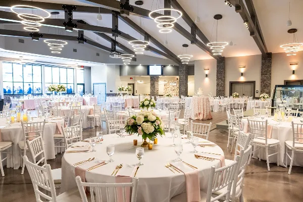 Wedding reception hall with round tables, white chairs, floral centerpieces, and a wedding cake under modern ring lighting.