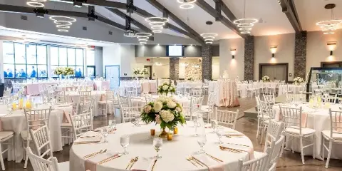 Wedding reception hall with round tables, white chairs, floral centerpieces, and a wedding cake under modern ring lighting.