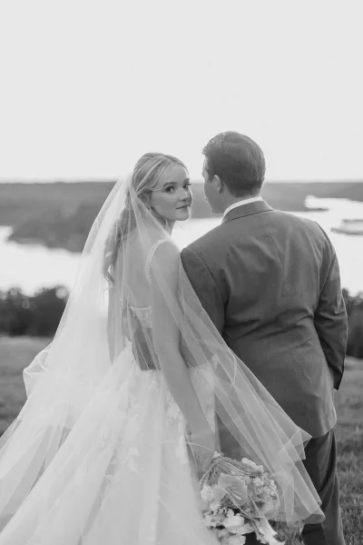 A black and white photograph of a bride and groom standing together outdoors, with the bride looking over her shoulder at the camera.