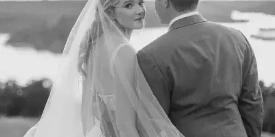 A black and white photograph of a bride and groom standing together outdoors, with the bride looking over her shoulder at the camera.