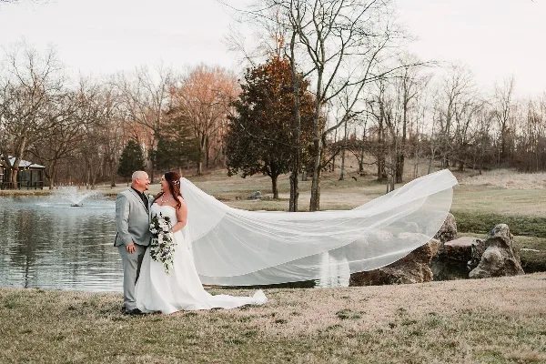 A bride and groom standing by a lake with the bride's long veil blowing in the wind.