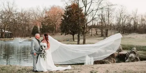 A bride and groom standing by a lake with the bride's long veil blowing in the wind.