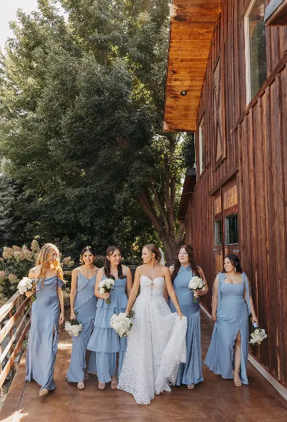 A bride and bridesmaids in blue dresses walking along a wooden deck next to a rustic barn structure.