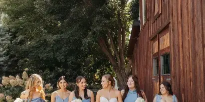 A bride and bridesmaids in blue dresses walking along a wooden deck next to a rustic barn structure.