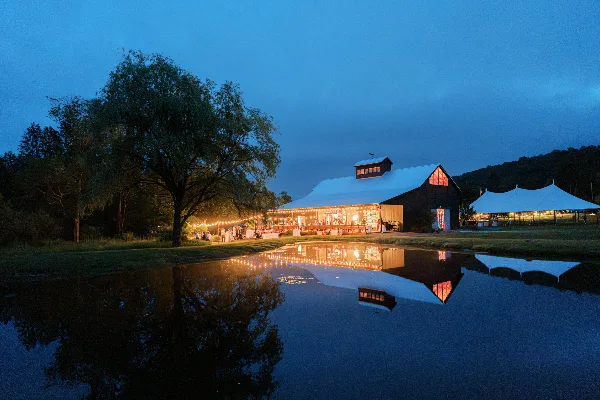 A wedding venue with a white reception tent and string lights reflected in a pond at dusk.