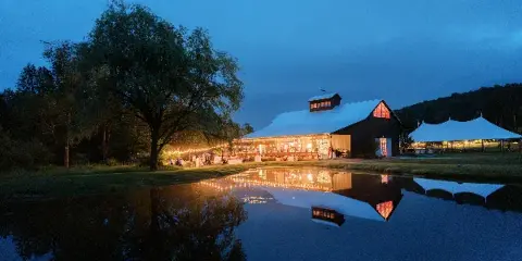 A wedding venue with a white reception tent and string lights reflected in a pond at dusk.