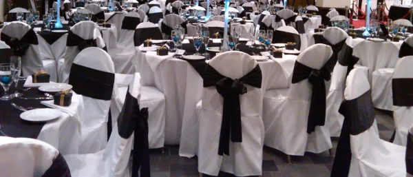 Banquet chairs with white covers and black sashes arranged around tables with tablecloths and place settings.