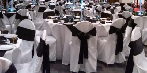 Banquet chairs with white covers and black sashes arranged around tables with tablecloths and place settings.