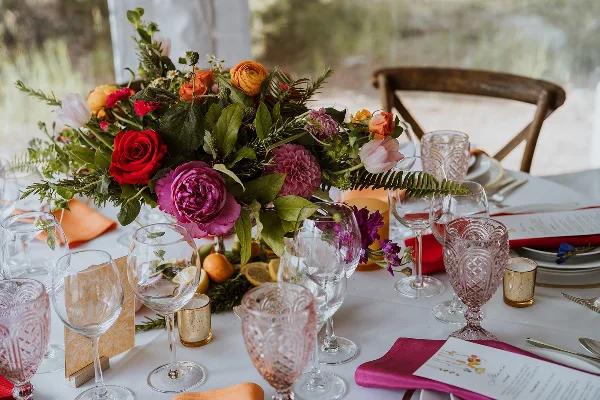 A vibrant wedding table centerpiece featuring red roses, pink peonies, and greenery, surrounded by glassware and place settings.