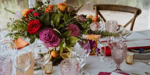 A vibrant wedding table centerpiece featuring red roses, pink peonies, and greenery, surrounded by glassware and place settings.