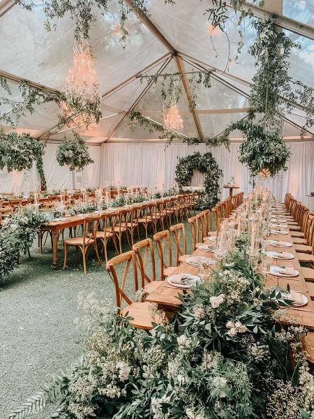 A long wooden dining table set with white plates and glassware, surrounded by wooden chairs and lush floral centerpieces under a clear tent with hanging greenery and chandeliers.