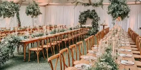 A long wooden dining table set with white plates and glassware, surrounded by wooden chairs and lush floral centerpieces under a clear tent with hanging greenery and chandeliers.