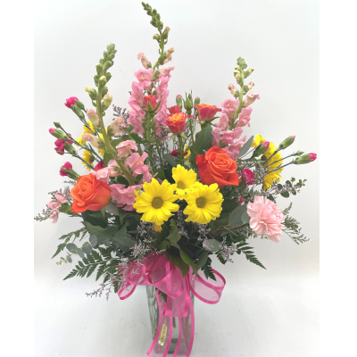 A vibrant floral arrangement in a clear vase featuring pink snapdragons, orange roses, yellow chrysanthemums, and greenery, tied with a pink ribbon.