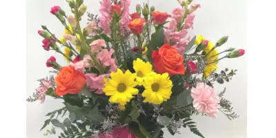 A vibrant floral arrangement in a clear vase featuring pink snapdragons, orange roses, yellow chrysanthemums, and greenery, tied with a pink ribbon.