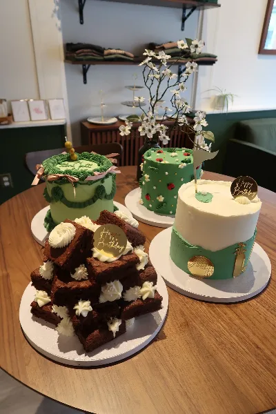 A display of four custom cakes and a stack of brownies on a wooden table, featuring green and white frosting designs.