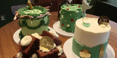 A display of four custom cakes and a stack of brownies on a wooden table, featuring green and white frosting designs.