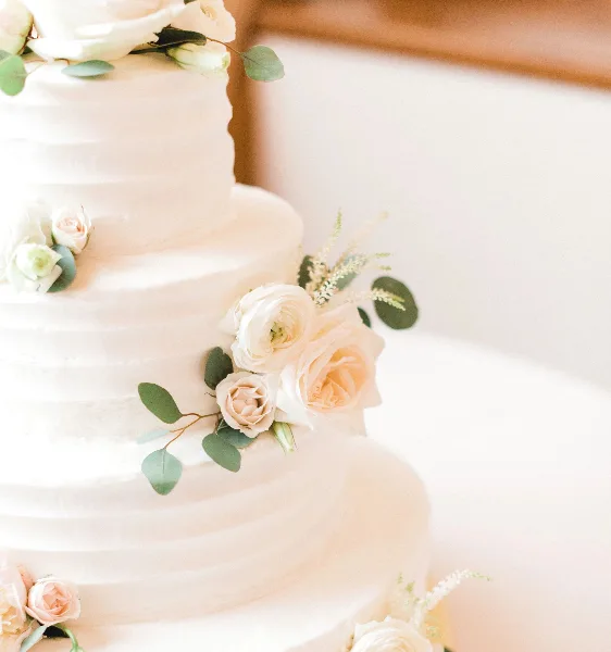 A close-up of a white, multi-tiered wedding cake decorated with white and blush roses and eucalyptus leaves.