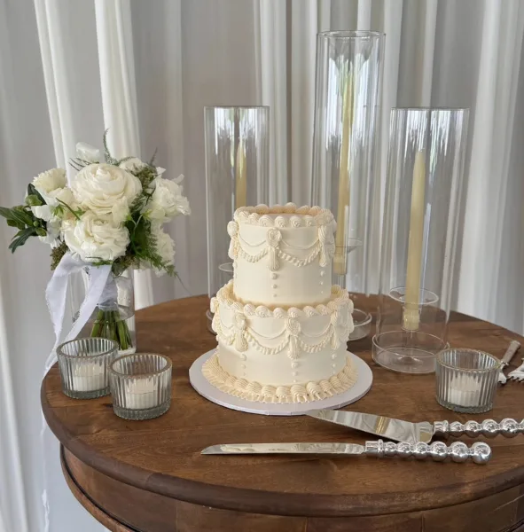 A two-tier white wedding cake with decorative buttercream swags and piping, displayed on a wooden table with a white floral bouquet and tall glass candle holders.