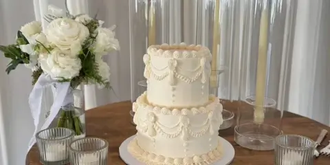 A two-tier white wedding cake with decorative buttercream swags and piping, displayed on a wooden table with a white floral bouquet and tall glass candle holders.