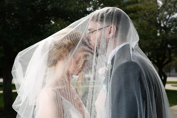 A bride and groom standing close together under a white veil in an outdoor setting.