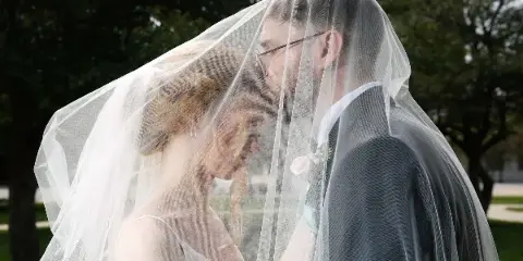 A bride and groom standing close together under a white veil in an outdoor setting.