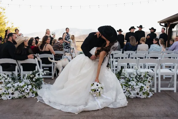 Bride and groom kiss during an outdoor wedding ceremony with seated guests and white floral arrangements.