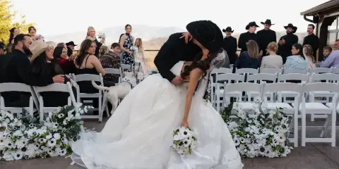 Bride and groom kiss during an outdoor wedding ceremony with seated guests and white floral arrangements.