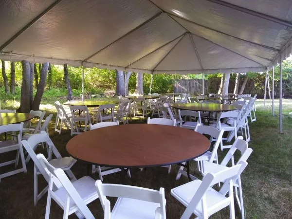 An outdoor event setup featuring a large white tent, round tables, and white folding chairs arranged on a grassy lawn.