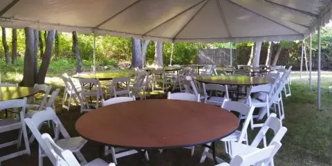 An outdoor event setup featuring a large white tent, round tables, and white folding chairs arranged on a grassy lawn.