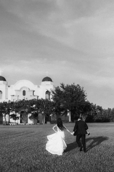 A black and white photo of a bride and groom running hand-in-hand across a lawn with a church in the background.