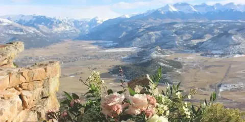 A floral arrangement with pink and white roses and greenery placed on a stone ledge overlooking a mountain valley.
