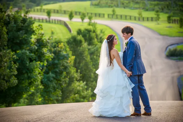 A bride in a white ruffled gown and a groom in a blue suit standing together outdoors on a paved overlook with green hills in the background.