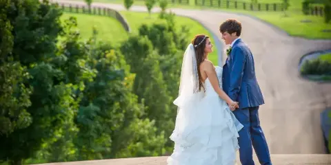 A bride in a white ruffled gown and a groom in a blue suit standing together outdoors on a paved overlook with green hills in the background.