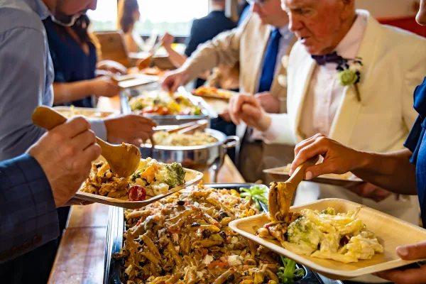 Catering staff serving a buffet of pasta and vegetable dishes to guests at a formal event.