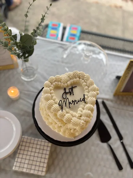 A heart-shaped white buttercream cake with piped rosettes and 'Just Married' written in black calligraphy, displayed on a table with a cake server and floral decor.