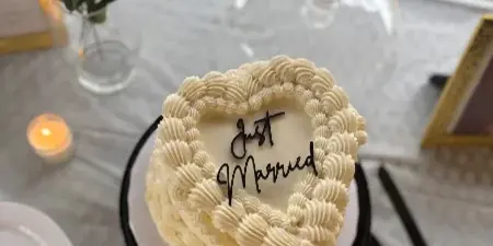 A heart-shaped white buttercream cake with piped rosettes and 'Just Married' written in black calligraphy, displayed on a table with a cake server and floral decor.